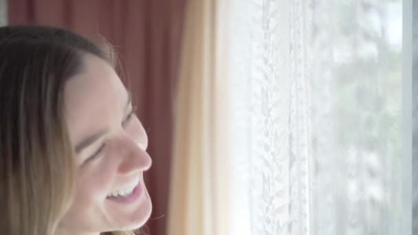Šťastná dívka se směje při pohledu ven oknem se závěsy, zblízka - Záběry, video