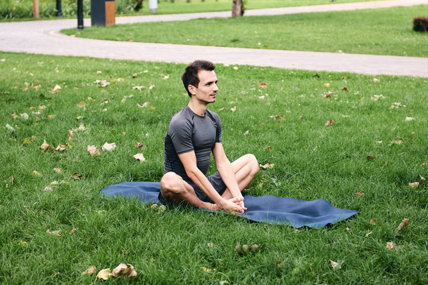 Fiatal férfi sportruházatban jógázik a parkban. Gyakorolja az ászanát a szabadban. Zöld füvön gyakorolni a jóga szőnyegen. Férfi ül pillangó lótusz pózol, nyugodt és meditáció.Fitness és az egészséges életmód - Fotó, kép