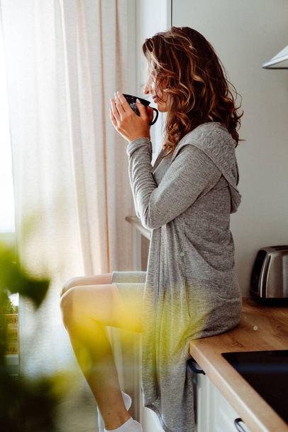 Young pretty woman drinks coffee sitting at her kitchen at home and looking out the window enjoys of morning - Photo, image