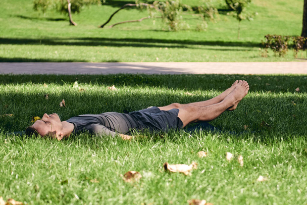 Young athletic man in sportswear doing yoga in the park. Practice asana outdoors. People exercising stretching on green grass with yoga mat. Fitness and healthy lifestyle. Concept of calm and meditation. - Foto, imagen