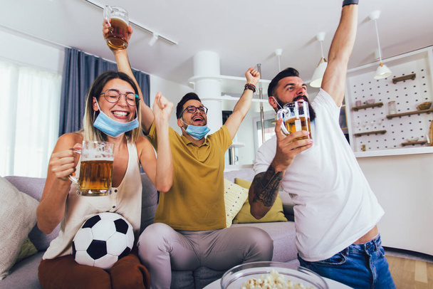 Gruppe junger Freunde schaut sich zu Hause ein Fußballspiel an, jubelt und trinkt Bier, trägt Schutzmaske. - Foto, Bild