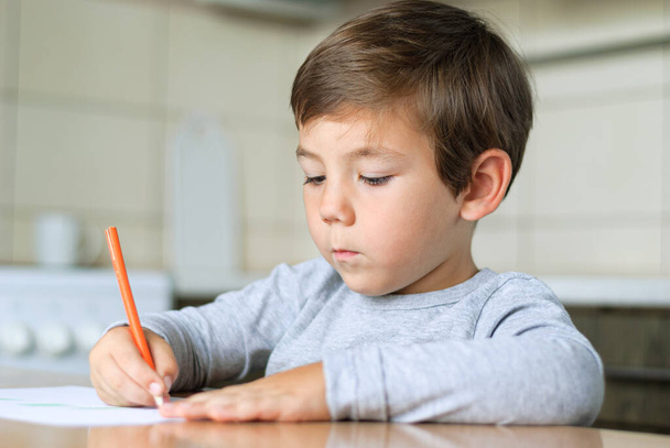 Junge in der Küche am Tisch zeichnet mit Bleistift auf ein Blatt Papier - Foto, Bild