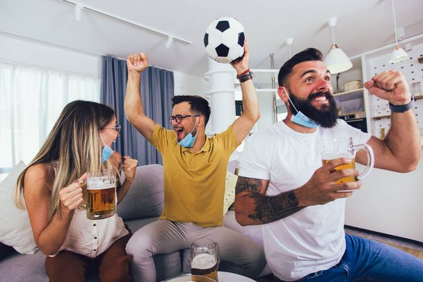 Grupo de jóvenes amigos viendo un partido de fútbol en casa, animando y bebiendo cerveza, usando máscara protectora. - Foto, Imagen