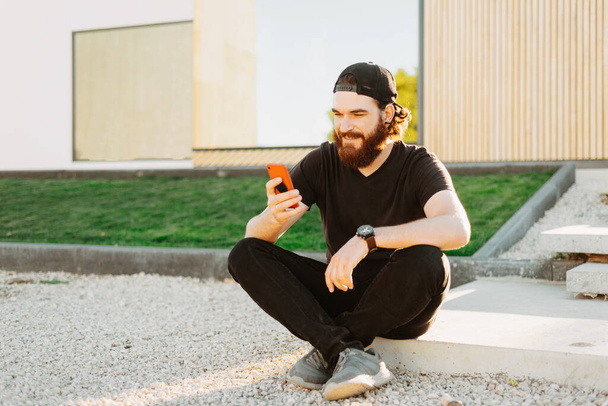 Фото молодого бородатого чоловіка, який сидить перед своїм будинком і використовує смартфон і посміхається
 - Фото, зображення