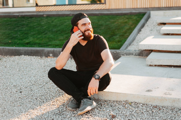 happy bearded hipster man sitting outdoor and talking on smartphone near his house - Photo, Image