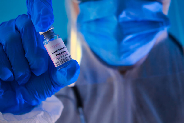 Доктор в голубых перчатках, маске, очках, стетоскопе, держа розовый ковид вакцины 19 бутылка, передний план. - Фото, изображение