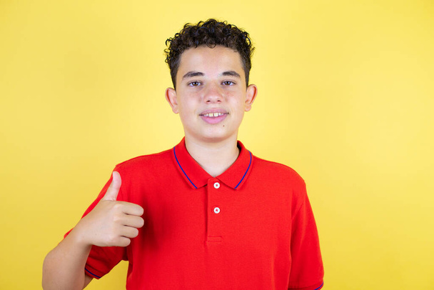 Красивый мальчик-подросток на изолированном желтом фоне улыбается и делает сигнал ОК большим пальцем - Фото, изображение