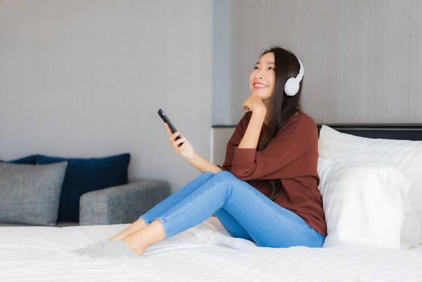 Πορτρέτο όμορφη νεαρή ασιατική γυναίκα χρησιμοποιούν έξυπνο κινητό τηλέφωνο με ακουστικά για να ακούσετε μουσική στο κρεβάτι στο εσωτερικό του υπνοδωματίου - Φωτογραφία, εικόνα