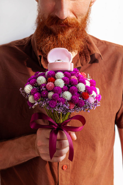 Marriage proposal. Handsome elegant man in brown shirt gives white gold ring in pink box beautiful flowers bouquet. Bearded guy proposing girlfriend to marry him. Romantic date, engagement celebration - Foto, afbeelding