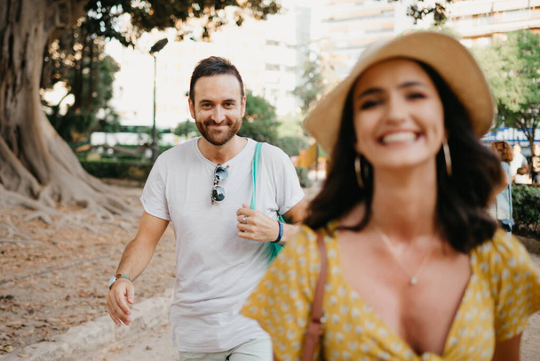 A smiling brunette wears a hat and a yellow dress goes before her boyfriend with a beard near an old Valencian Ficus Macrophylla tree in Spain. A couple of tourists enjoying Valencia in the evening. - Photo, Image