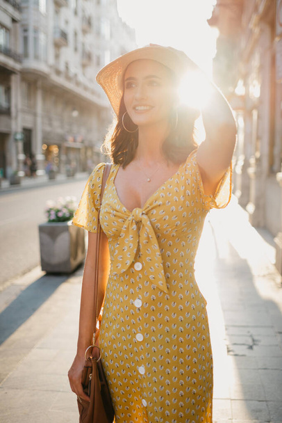 Μια όμορφη νεαρή γυναίκα με ένα κίτρινο φόρεμα με βαθύ ντεκολτέ κρατά ένα καπέλο στο κεφάλι της στο ηλιοβασίλεμα στην Ισπανία. Ένα πανέμορφο χαμογελαστό κορίτσι στέκεται σε ένα παλιό δρόμο στη Βαλένθια το βράδυ.  - Φωτογραφία, εικόνα