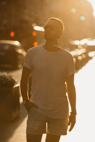 Un homme en lunettes de soleil, un t-shirt blanc et un short font du tourisme à Valence dans les rayons du coucher de soleil sur une vieille rue. Un mec barbu profite de l'Espagne le soir. - Photo, image