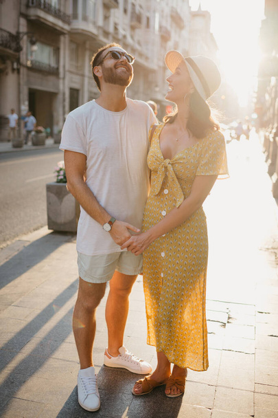Meisje met een hoed en een gele jurk met een vallende halslijn en haar vriendje met een baard lachen elkaar uit terwijl ze elkaars hand vasthouden in Spanje. Een paar geliefden knuffelen elkaar bij zonsondergang in Valencia - Foto, afbeelding