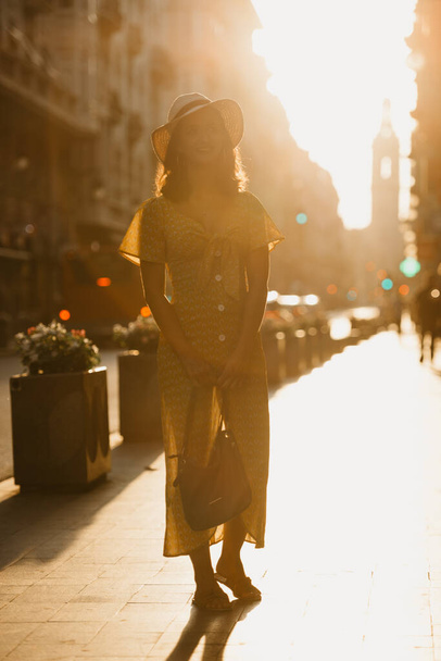 A cute young woman in a hat and a yellow dress with a plunging neckline is holding a leather bag on the sunset in Spain. A gorgeous smiling girl is posing on an old street in Valencia in the evening. - Photo, Image