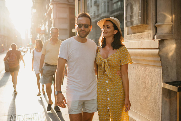 Um homem com barba e óculos de sol abraça a cintura com a mão de uma namorada de chapéu e um vestido amarelo com um decote em Espanha. Um par de amantes no pôr do sol em Valência. - Foto, Imagem
