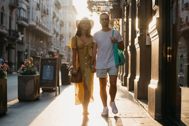 Tyttö hattu päässä ja keltainen mekko syvä pääntie ja hänen poikaystävänsä parta ja aurinkolasit kävelevät pitäen toisiaan kädestä Espanjassa. Pari turistia auringonlaskussa Valenciassa - Valokuva, kuva