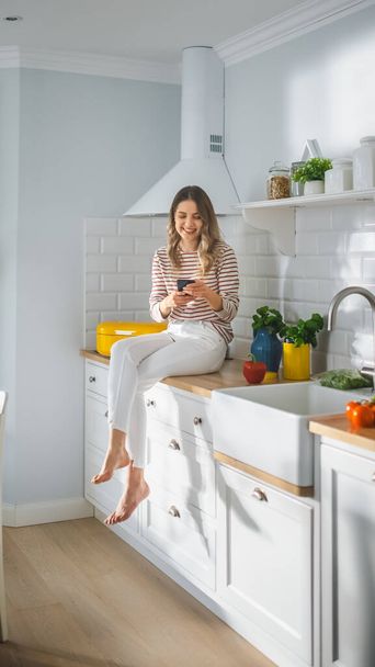 Belle jeune femme en pull rayé et pantalon blanc est assis sur un mobilier de cuisine et en utilisant son smartphone dans une cuisine moderne ensoleillée. Elle est heureuse et excitée tout en naviguant sur Internet. - Photo, image