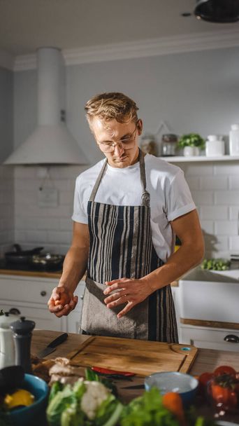 Handsome Man in White Shirt and Apron is Making a Healthy Organic Salad Meal in a Modern Sunny Kitchen. Hipster Man in Glasses Cooking. Natural Clean Diet and Healthy Way of Life Concept. - 写真・画像