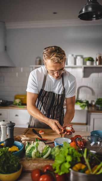 Handsome Man in White Shirt and Apron is Making a Healthy Organic Salad Meal in a Modern Sunny Kitchen. Hipster Man in Glasses Cooking. Natural Clean Diet and Healthy Way of Life Concept. - 写真・画像