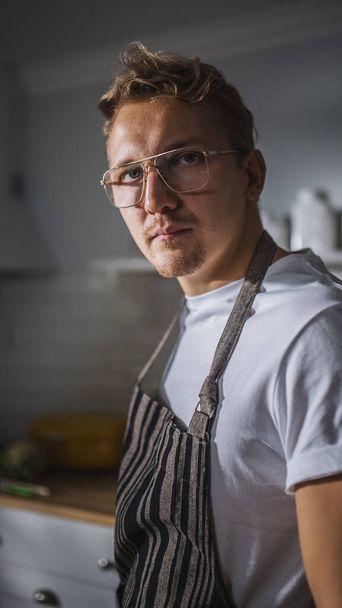Portrait Shot of a Handsome Cook in White Shirt and Apron Posing on Camera in a Bright Modern Kitchen. Hipster Man in Glasses. Natural Clean Diet, Healthy Way of Life Concept - Fotografie, Obrázek
