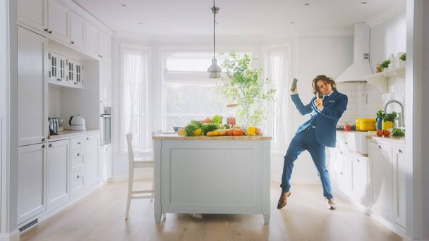 Happy Young Man with Long Hair Dancing at Home while Wearing Blue Business Suit. He is Listening to Music on a Mobile. Energetic Man Using Smartphone in a Kitchen with Healthy Vegetables. - Фото, изображение