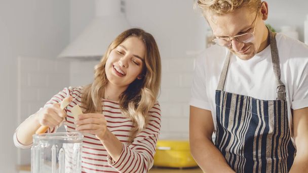 Handsome Young Man in Glasses Wearing Apron and Beautiful Girl are Making A Smoothie in the Kitchen. Happy Couple are Preparing Healthy Organic Beverage. Male and Female at Home on a Sunny Day. - Foto, imagen