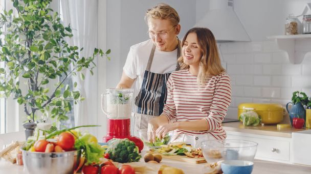 Handsome Young Man in Glasses Wearing Apron and Beautiful Girl are Making A Smoothie in the Kitchen. Happy Couple are Preparing Healthy Organic Beverage. Male and Female at Home on a Sunny Day. - Photo, image
