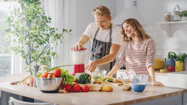 Handsome Young Man in Glasses Wearing Apron and Beautiful Girl are Making A Smoothie in the Kitchen. Happy Couple are Preparing Healthy Organic Beverage. Male and Female at Home on a Sunny Day. - Foto, immagini