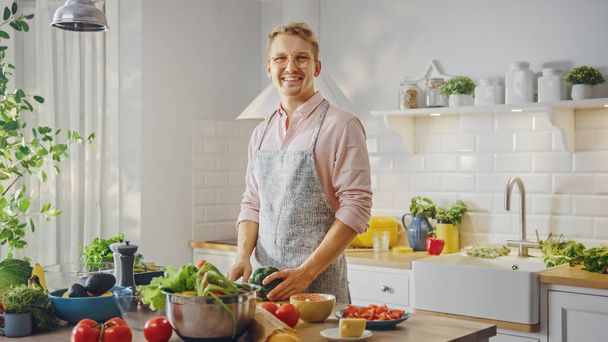 Handsome Man in Pink Shirt and Apron is Making a Healthy Organic Salad Meal in a Modern Sunny Kitchen. Hipster Man in Glasses Smiles at the Camera. Natural Clean Diet and Healthy Way of Life Concept. - 写真・画像