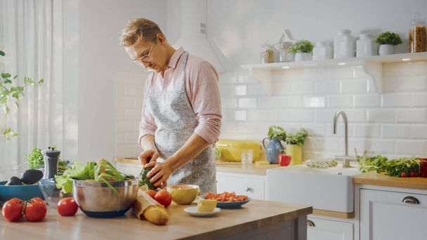Handsome Man in Pink Shirt and Apron is Making a Healthy Organic Salad Meal in a Modern Sunny Kitchen. Hipster Man in Glasses. Natural Clean Diet and Healthy Way of Life Concept. - 写真・画像