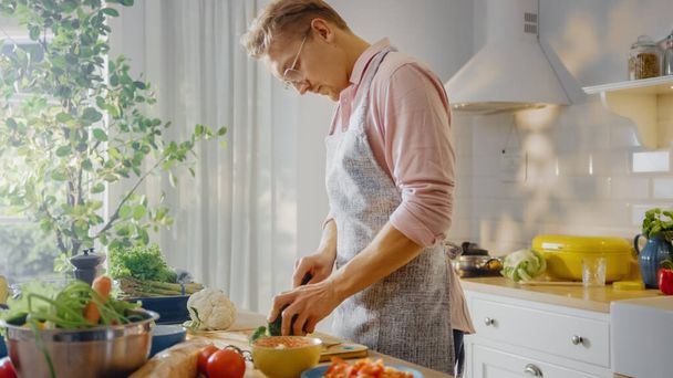 Homem bonito em camisa rosa e avental está fazendo uma refeição saudável salada orgânica em uma cozinha moderna ensolarada. Hipster Man in Glasses. Dieta Limpa Natural e Conceito de Modo de Vida Saudável. - Foto, Imagem