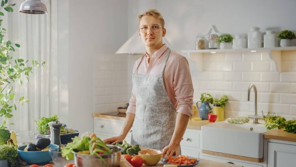 Handsome Man in Pink Shirt and Apron is Making a Healthy Organic Salad Meal in a Modern Sunny Kitchen. Hipster Man in Glasses Looking at the Camera. Natural Clean Diet and Healthy Way of Life Concept. - Fotó, kép