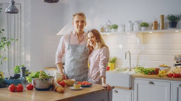眼鏡をかけてハンサムな若い男4月と美しい女の子を着ているキッチンでサラダを準備しています。幸せなカップルはお互いに抱き合っている。自然食と健康的なライフスタイルのコンセプト. - 写真・画像