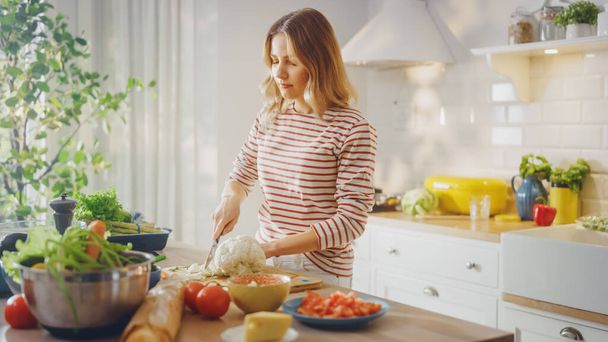 Young Female in Striped Jumper is Making a Healthy Organic Salad in a Modern Sunny Kitchen. Authentic Woman is Chopping a Cauliflower with a Knife. Natural Clean Diet and Healthy Way of Life Concept. - Foto, afbeelding