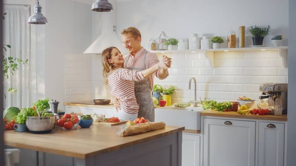 Handsome Young Man in Pink Shirt and Apron and Beautiful Girl in Striped Jumper are Creatively Dancing in the Kitchen. Sunny Modern Kitchen with Healthy Green Vegetables on Table. Happy Couple at Home - Photo, image