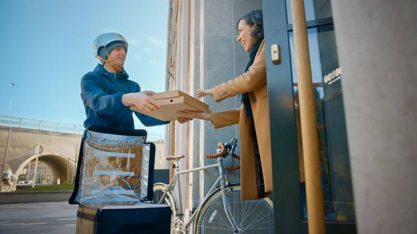 Happy Food Delivery Homme portant un sac à dos thermique sur un vélo livre la commande de pizza à une belle cliente. Un messager livre un déjeuner à emporter à une magnifique fille dans un immeuble de bureaux de district moderne - Photo, image
