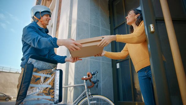 Happy Food Delivery Man Wearing Thermal Backpack on a Bike Delivers Pizza Order to a Beautiful Female Customer. Courier Delivers Takeaway Fast Food to a Smiling Girl in Office Building. Low Angle - Photo, image