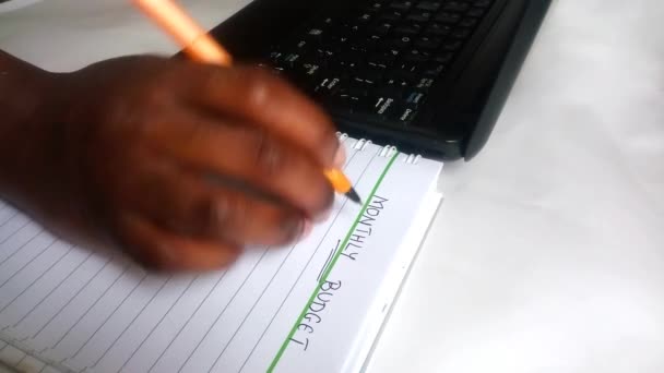 Ежемесячный бюджет написан на блокноте. Африканская леди держит ручку и с тревогой стучит и суетится, гадая, что написать. Ноутбук в фоновом режиме. Домашний офис. Чёрный. Этничность, разнообразие. - Кадры, видео