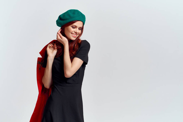 A woman holds in her hand a red coat and a dark T-shirt Green hat cropped view of a light background - 写真・画像