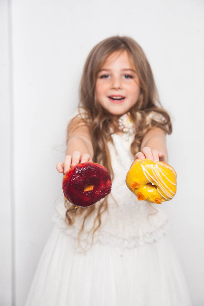 Little happy cute preschool girl is eating colourful donuts on white background isolated. Emotional child is having fun with sweets. Tasty sweet food for kids. Free copy space for text mockup  - Photo, Image