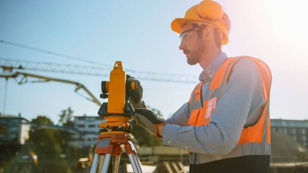 Construction Worker Using Theodolite Surveying Optical Instrument for Measuring Angles in Horizontal and Vertical Planes on Construction Site. Worker in Hard Hat Making Projections for the Building. - Photo, image