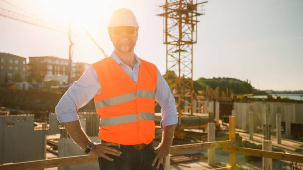 Confident Bearded Head Civil Engineer-Architect in Sunglasses is Smiling on Camera in a Construction Site on a Sunny Bright Day. Man is Wearing a Hard Hat, Shirt, Jeans and an Orange Safety Vest.  - Fotoğraf, Görsel