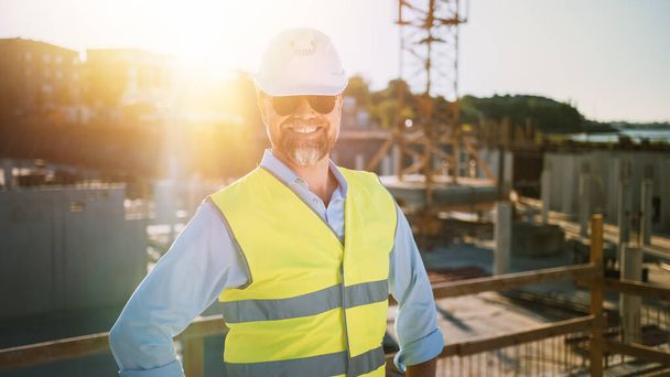 Confident Bearded Head Civil Engineer-Architect in Sunglasses is Smiling on Camera in a Construction Site on a Sunny Bright Day. Man is Wearing a Hard Hat, Shirt, Jeans and a Yellow Safety Vest.  - Zdjęcie, obraz
