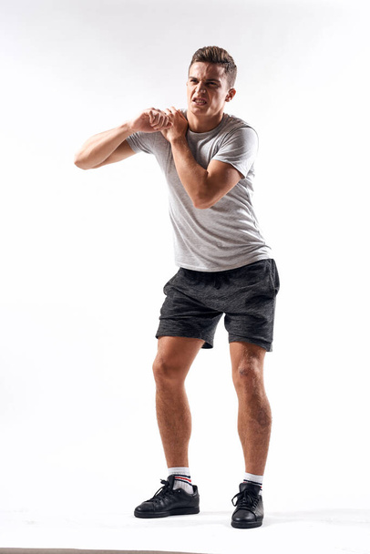 A sports man in shorts and a full-length T-shirt does exercises on a light background - Foto, imagen