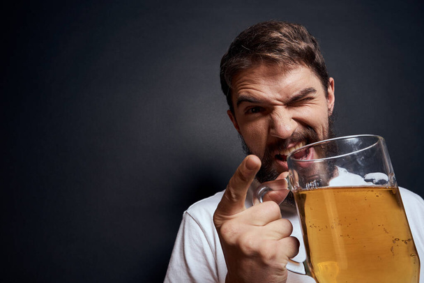 man with a mug of beer in a white t-shirt emotions lifestyle drunk on a dark isolated background - Photo, Image