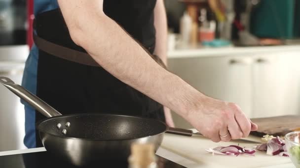 professionelle junge Koch in der heimischen Küche wirft dünne Scheiben Zwiebeln in eine Pfanne mit Öl. Zubereitung eines gebratenen Gerichts zum Mittag- oder Abendessen. Junk Food. Arbeit in der Küche in der Wohnung - Filmmaterial, Video
