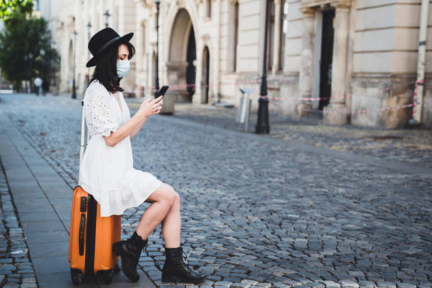 Seyahat çantasında oturan koruyucu maskeli güzel bayan turistin yan görüntüsü. Şapkalı ve beyaz elbiseli moda kadın gezgin cep telefonunda kontrol ve daktilo ediyor. Salgın Seyahat Konsepti - Fotoğraf, Görsel