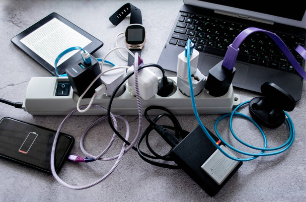 Muchos gadgets: smartphone, auriculares inalámbricos, portátil, reloj inteligente, batería de la cámara y e-book se están cargando en una tira de alimentación. - Foto, imagen
