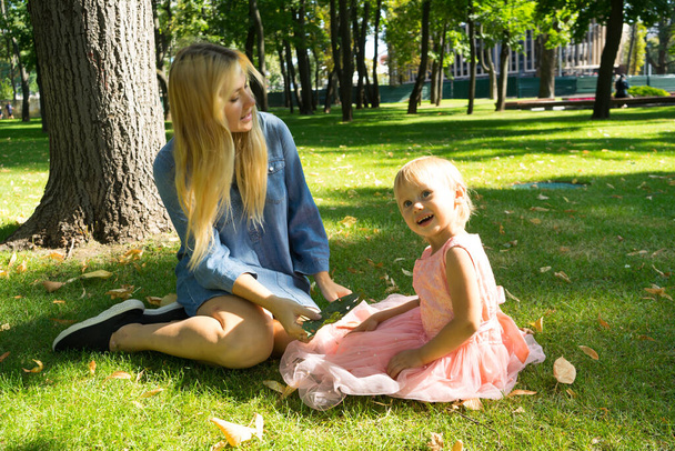 Beautiful mother and daughter with blonde hair playing together in the sunny park .Mother and daughter sitting together on green grass.Mother and daughter having funny conversation.Happy family concep - Valokuva, kuva