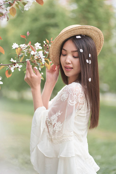 bela mulher asiática em um vestido branco e chapéu de palha, de pé perto de uma árvore com flores brancas e segurando ramos com as mãos, olhando para longe                                - Foto, Imagem